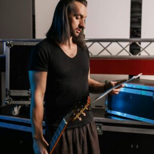 Male musician with long hair holding guitar and music sheet in a backstage setting.